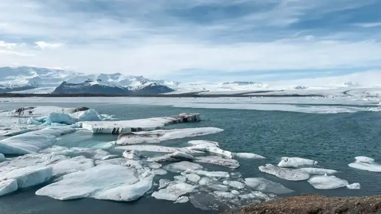 Jökulsárlón Glacier Lagoon in Iceland, featuring floating icebergs of various sizes drifting in the calm, blue waters. The background showcases snow-covered mountains and a vast glacier under a partly cloudy sky. The contrast between the icy white formations and the deep blue water creates a stunning and serene Arctic landscape.