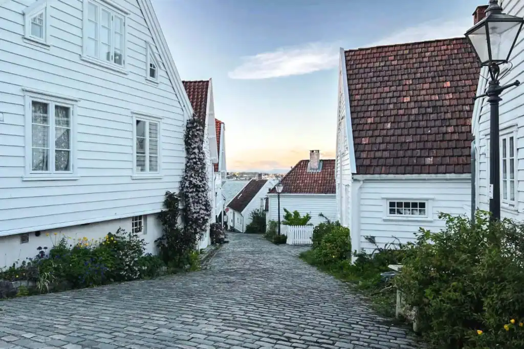 A charming cobblestone street in Stavanger, Norway, lined with traditional white wooden houses with red-tiled roofs. Greenery and colorful flowers grow along the walls, adding a touch of warmth to the scene. The street slopes gently downward, leading to a view of the water and a pastel-hued sunset in the distance.