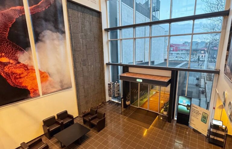 The modern lobby of Hotel Selfoss, Iceland, features high ceilings, floor-to-ceiling windows, and sleek black tiles reflecting the warm interior lighting. A striking volcanic eruption artwork covers one wall, complementing the dark stone accent feature beside it. The entrance leads to a glass-fronted exterior, where the town of Selfoss is visible through the large windows. Comfortable black leather armchairs and a coffee table create a welcoming seating area, adding to the stylish yet cosy ambience of the hotel.
