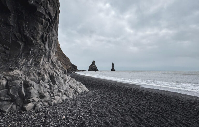 Dramatic basalt cliffs rise along the edge of Reynisfjara, Iceland’s famous black sand beach. The dark volcanic shoreline stretches toward the ocean, where powerful waves crash against the shore. In the distance, the Reynisdrangar sea stacks emerge from the misty waters, standing tall against the overcast sky. The moody atmosphere enhances the beach’s rugged and otherworldly beauty.