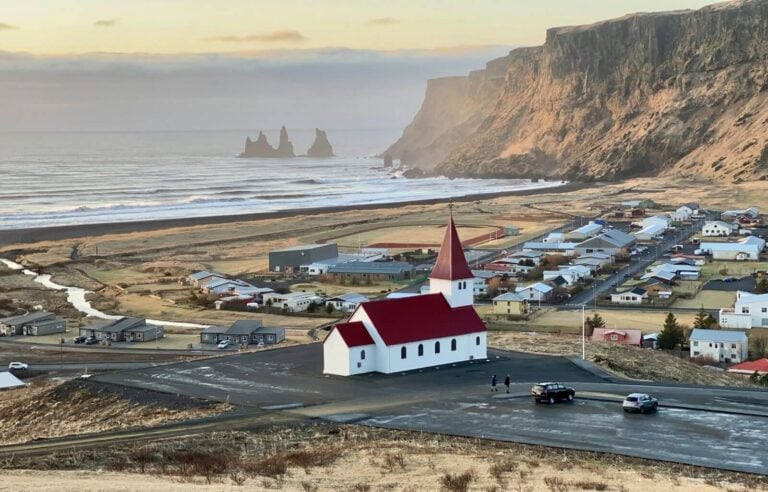 A white church with a red roof stands on a hill overlooking the town of Vík, Iceland. Below, the small coastal village is nestled between rugged cliffs and black sand beaches, with houses and buildings scattered across the landscape. In the distance, the Reynisdrangar sea stacks rise dramatically from the ocean as waves crash against the shore. The soft, golden light of the setting sun casts a warm glow over the town and surrounding mountains, creating a peaceful and picturesque scene.
