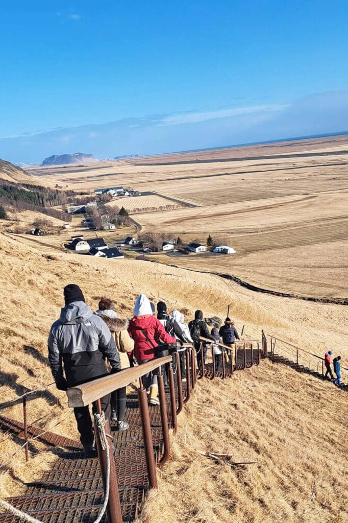 Group of hikers walking down a wooden staircase in a vast, open Icelandic landscape with rolling hills and distant mountains.