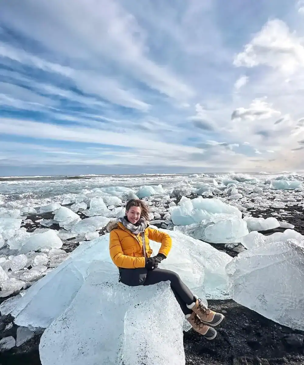 Woman in a mustard yellow winter jacket sitting on a large ice block on a black sand beach, surrounded by scattered glacier ice chunks beside the ocean. Dramatic streaked clouds stretch across the sky, creating a striking icy coastal landscape.