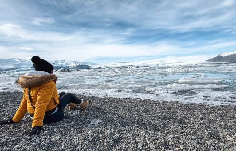 Person in yellow jacket and black hat sitting on rocky beach near icy water in Iceland.