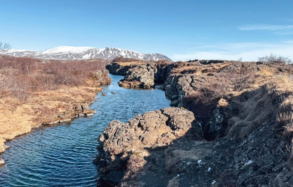 A river flowing through rocky terrain with snow-capped mountains in the background under a clear blue sky.
