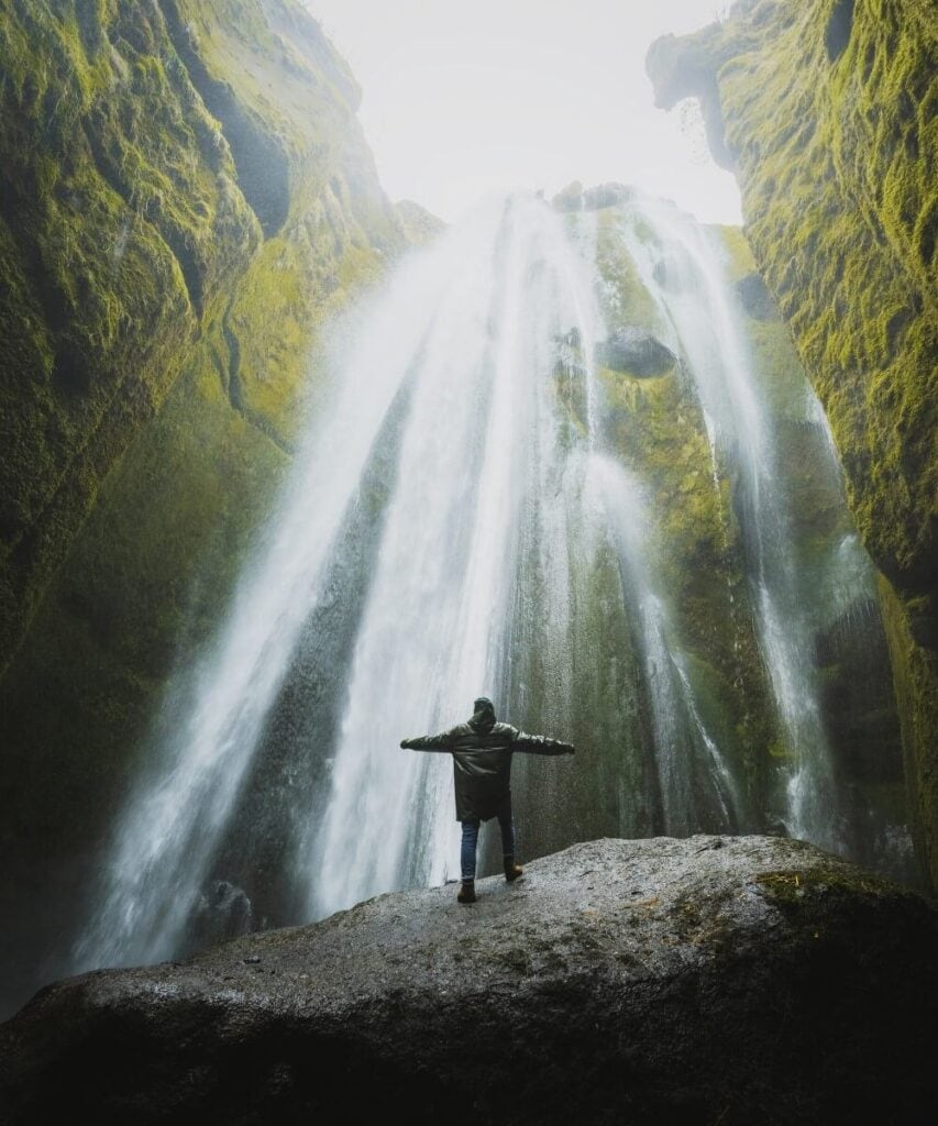 Person standing with arms outstretched in front of Seljalandsfoss waterfall surrounded by mossy cliffs.