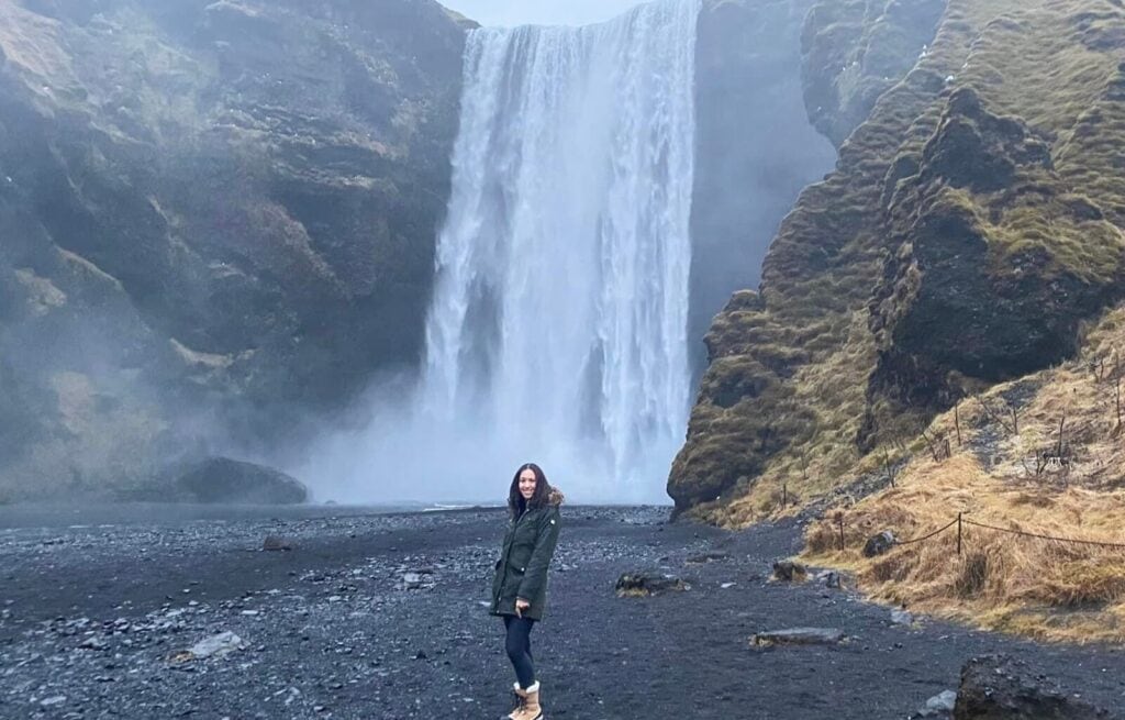 Woman standing near the base of Seljalandsfoss waterfall in Iceland with water cascading down cliffs behind her.