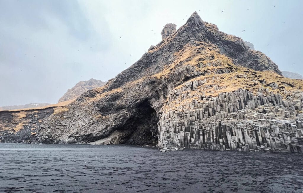 Cliffside with basalt columns and mountain in the background, near water, under cloudy sky.