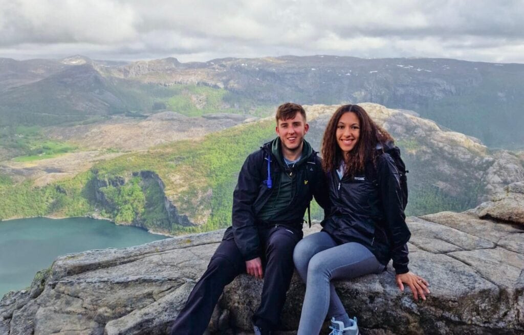 The founders of Under Northern Skies sit side by side on the edge of Pulpit Rock in Norway, smiling with a panoramic view of fjords, cliffs, and forested hills stretching out behind them under a cloudy sky.