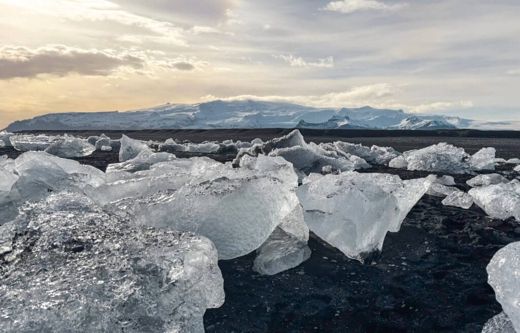 Ice chunks scattered across a black volcanic beach with distant mountains and a cloudy sky.