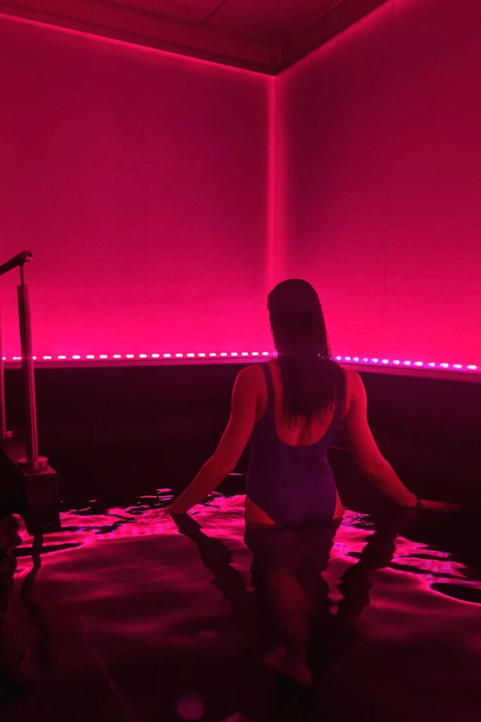 Woman relaxing in a pink-lit room with water, facing away from the camera.