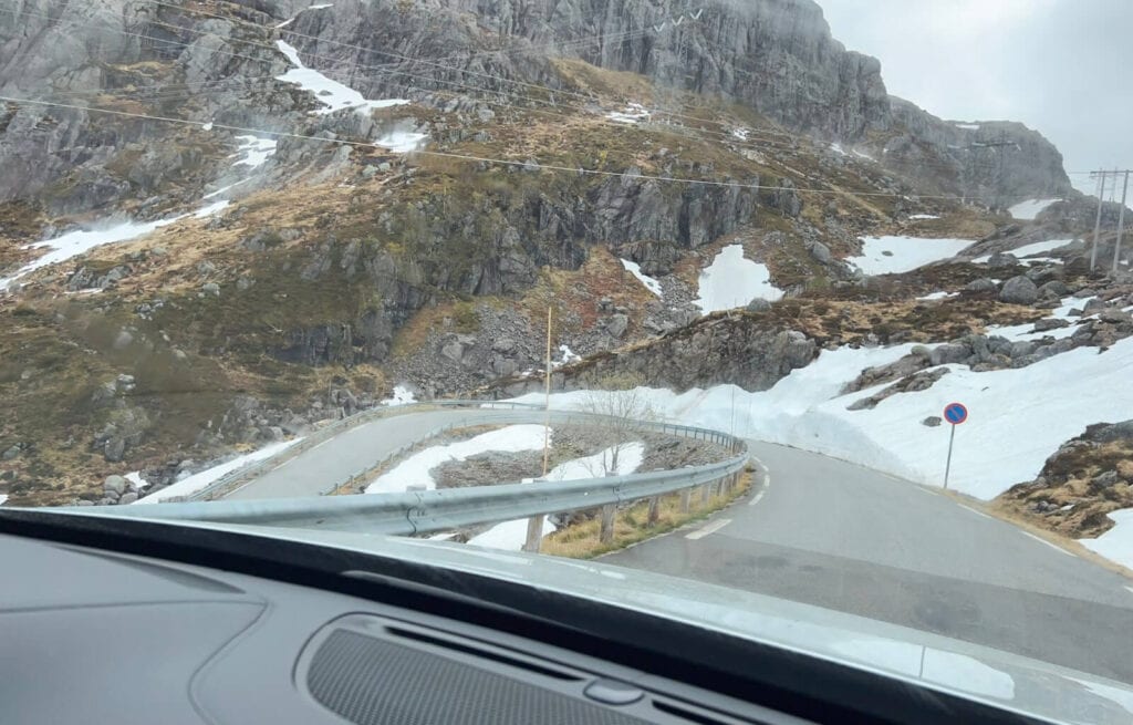 Winding mountain road with snow patches and rocky cliffs, seen from inside a car in a Nordic landscape.
