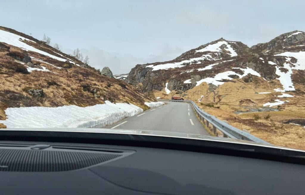 Scenic mountain road with snow patches and rocky slopes, viewed from inside a car in a Nordic landscape.