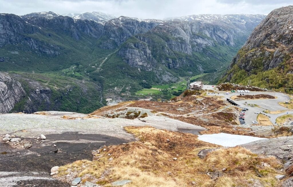Scenic view of a winding mountain road with parked cars, green valleys, and snow-capped peaks in the distance.