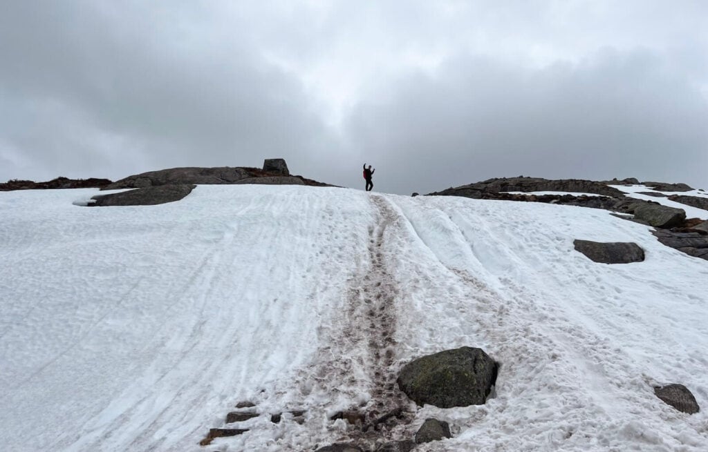 Hiker standing on snowy slope under cloudy sky, with rocky terrain and patches of snow.