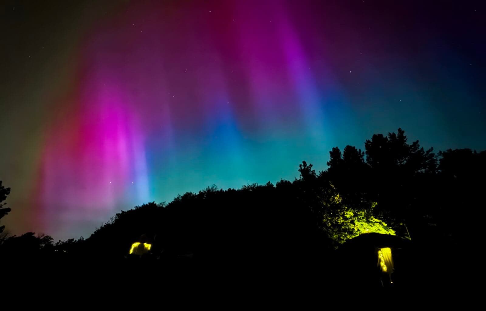 Vivid northern lights display with pink, green, and purple hues over silhouetted trees and small cabins.