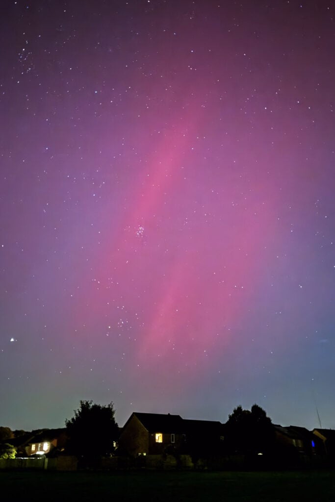 Vivid pink and purple aurora borealis streaks across a starry night sky over a rural landscape.
