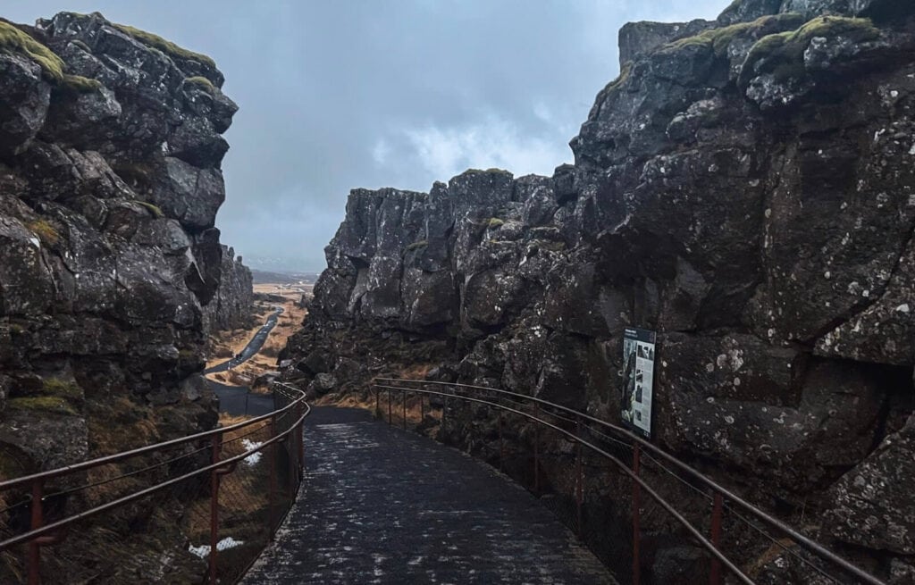 Narrow pathway between large dark volcanic rocks with metal railings on both sides, leading to a cloudy sky in the distance.