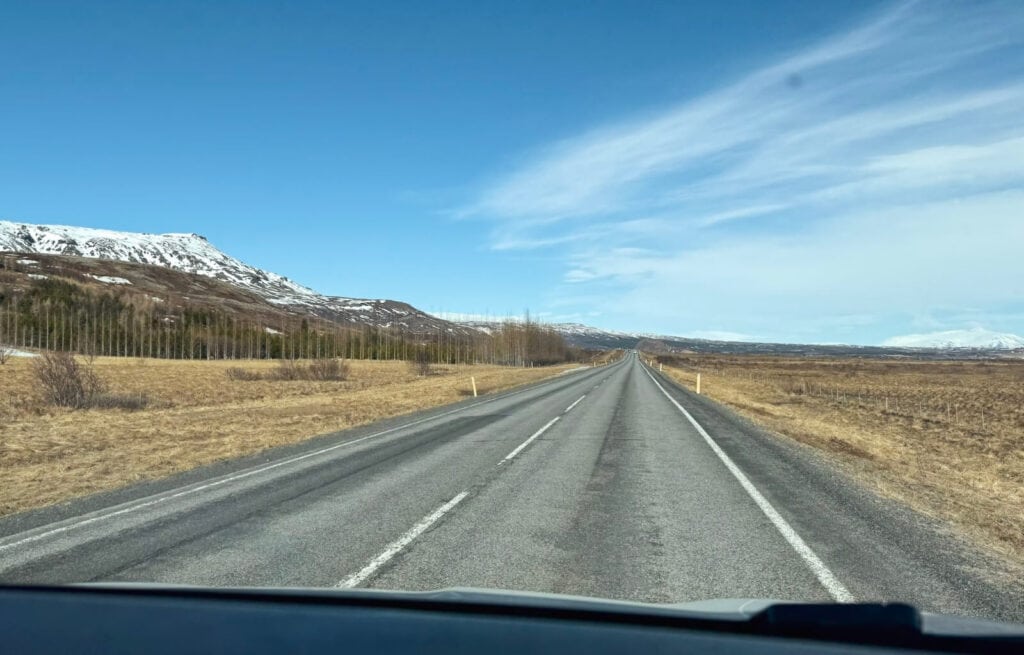 Long empty Icelandic road with mountains and snow in the distance under a blue sky.