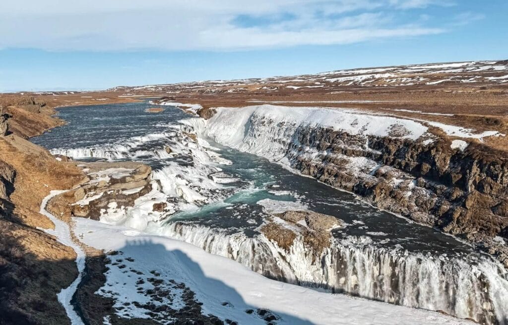 Gullfoss waterfall in winter with snow on the rocks and ice in the water, under a blue sky.