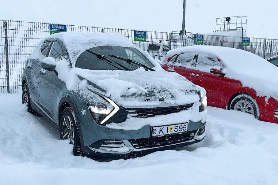 Snow-covered grey car parked in a lot with other vehicles, surrounded by snow and ice, under a grey sky.