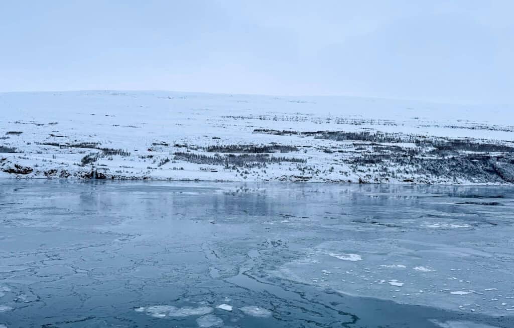 Frozen lake with cracked ice and snow-covered hills in the background under a cloudy sky.