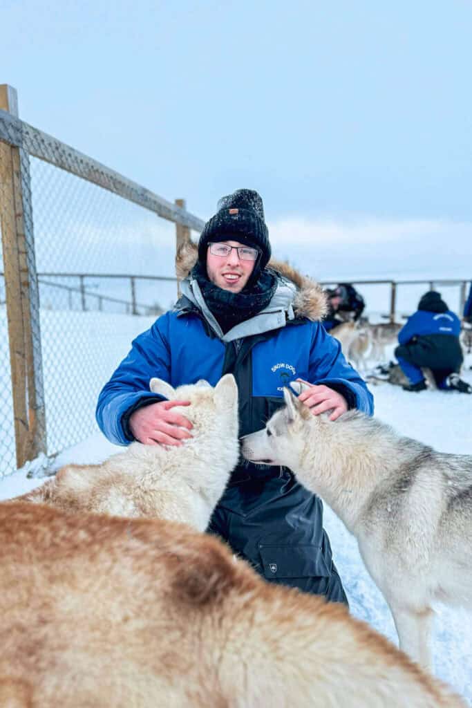 Person in blue winter gear with glasses petting two huskies in snowy enclosure.