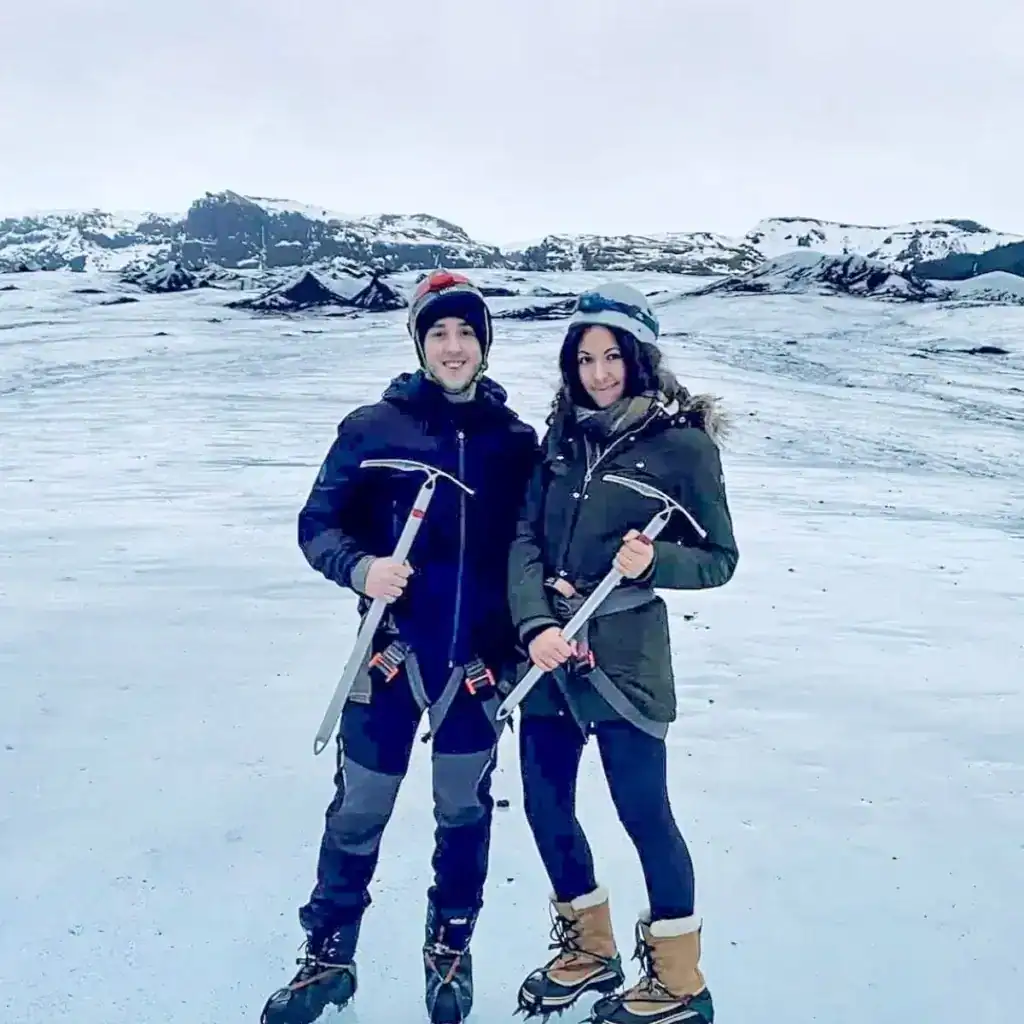 A smiling couple dressed in winter gear poses on a glacier, holding ice axes and wearing crampons on their boots. The man wears a black jacket, gray pants, and a beanie, while the woman wears a dark green parka with a fur-lined hood and a helmet. Snow-covered mountains and rugged ice formations stretch behind them under an overcast sky.
