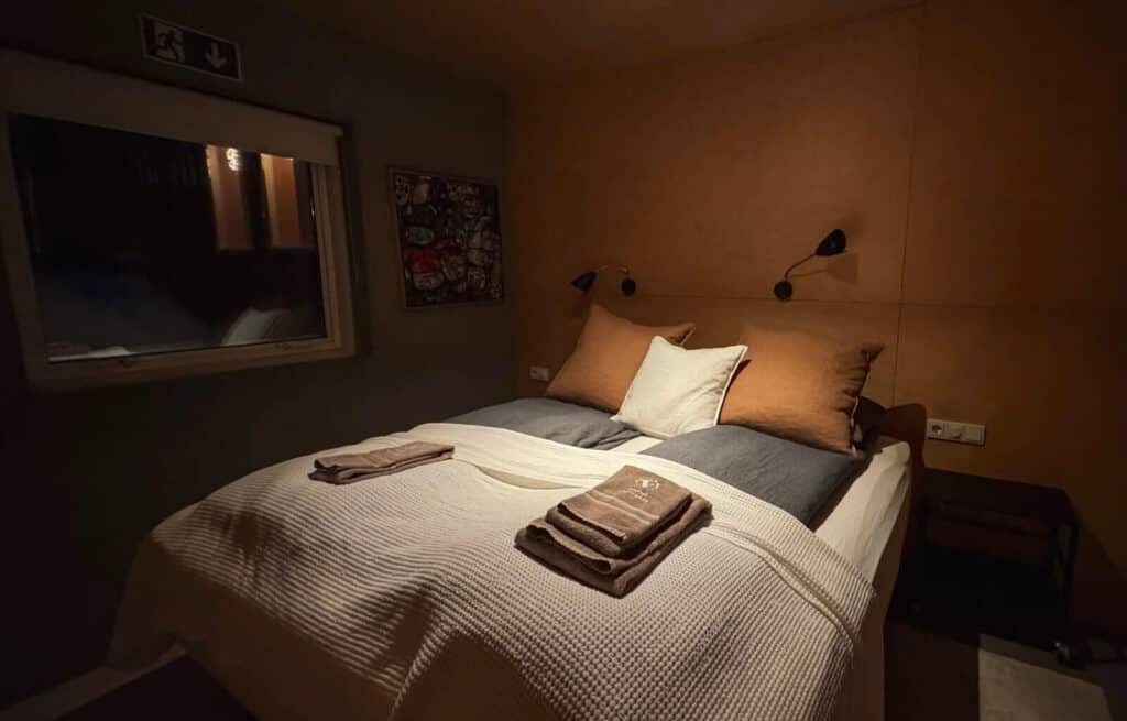 Comfortable bed with pillows and folded towels in a warmly lit bedroom with a window and artwork on the wall.
