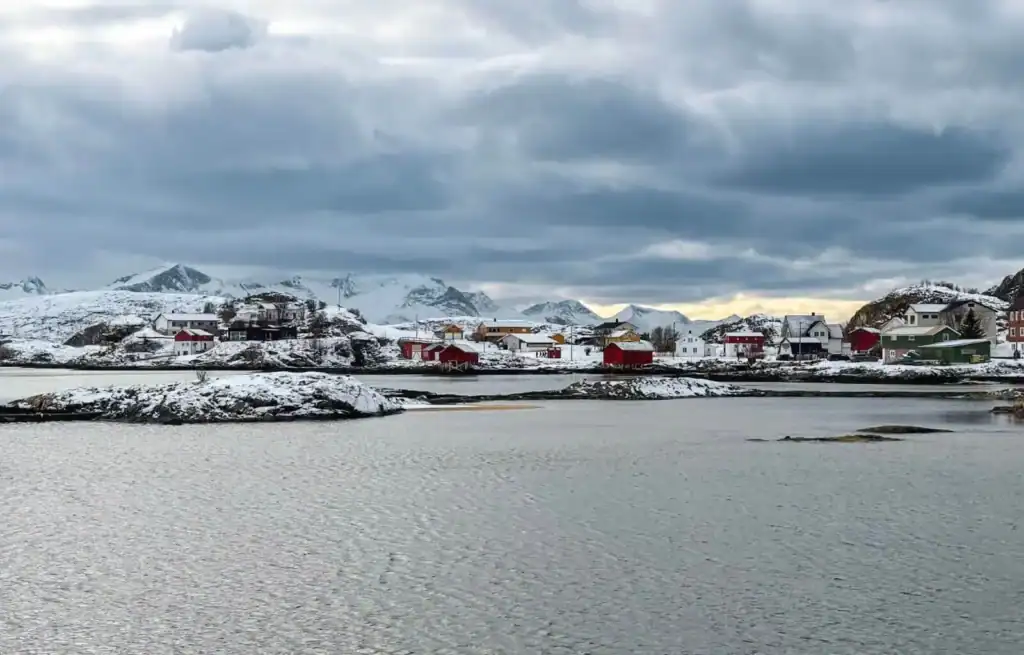 Snow-covered houses and mountains under cloudy sky in a Nordic coastal village.