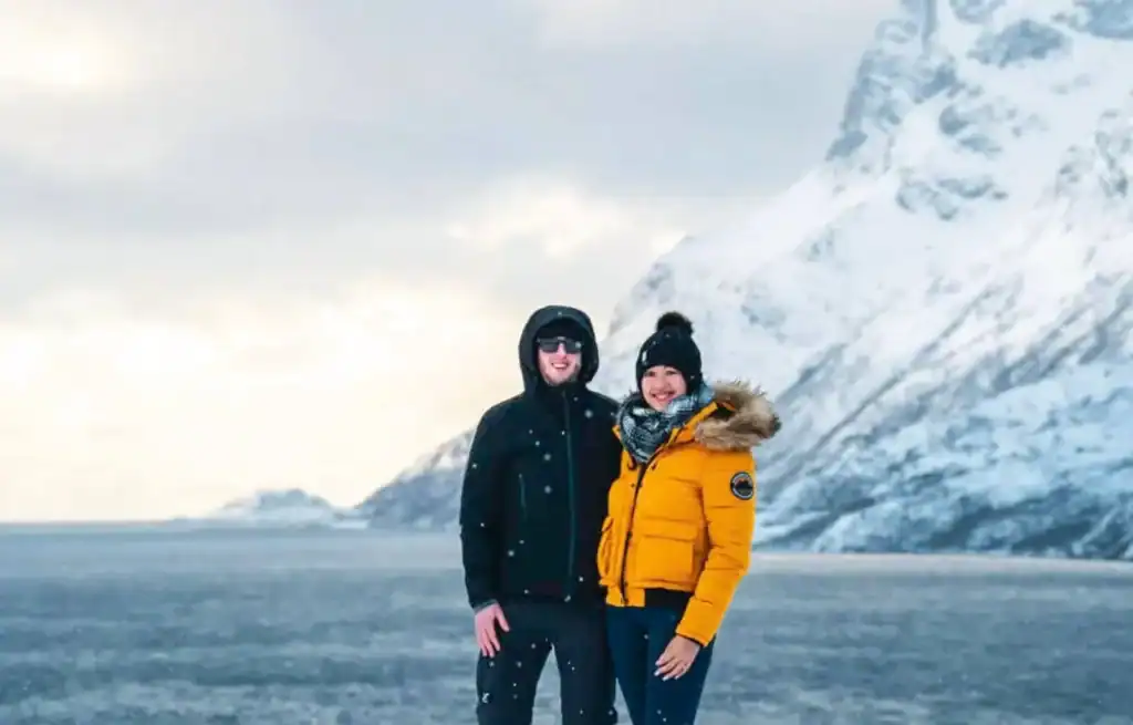 Two people in winter jackets stand on icy terrain with snow-covered mountains in the background.