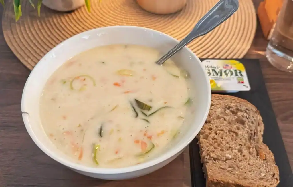 Bowl of creamy vegetable soup with a spoon, served with a slice of brown bread on a wooden table.