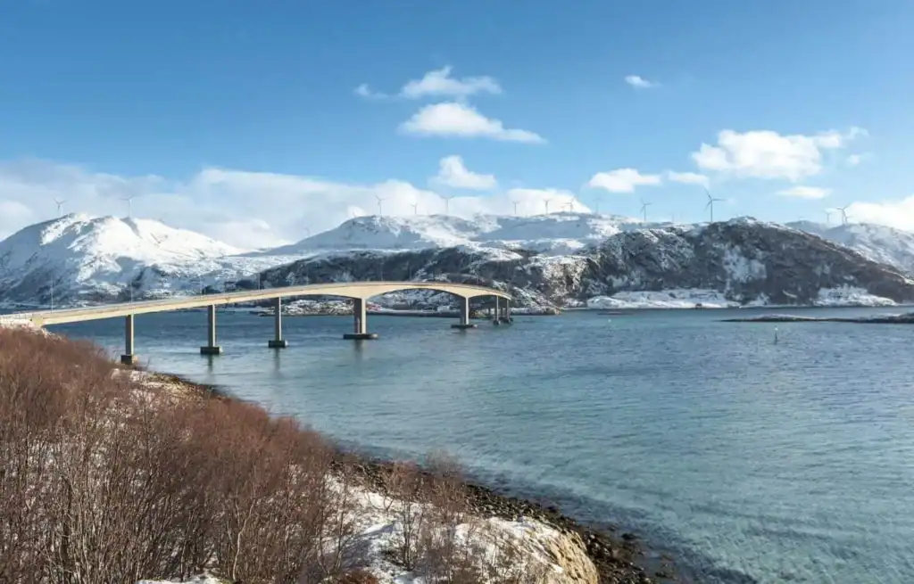 Snow-capped mountains and a bridge over calm water under a blue sky with clouds.