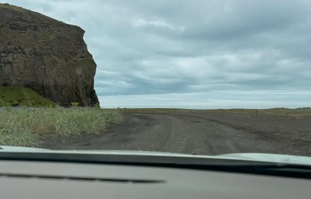Unpaved gravel road leading towards a cliff edge with cloudy sky overhead.