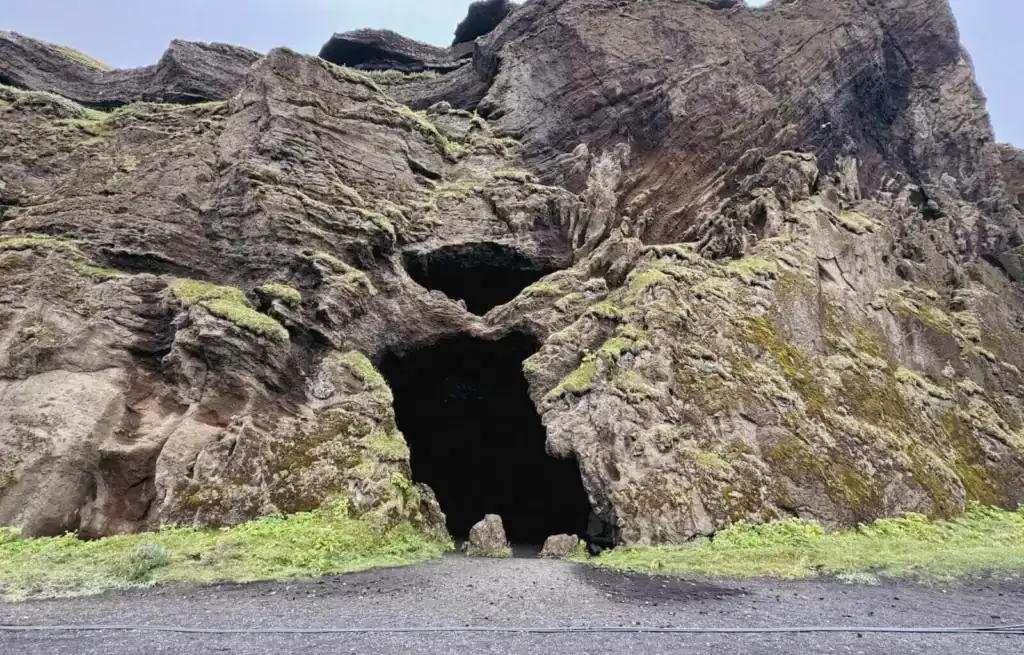 View of Yoda Cave with dark entrance in rugged volcanic rock formation, surrounded by green moss and grass.
