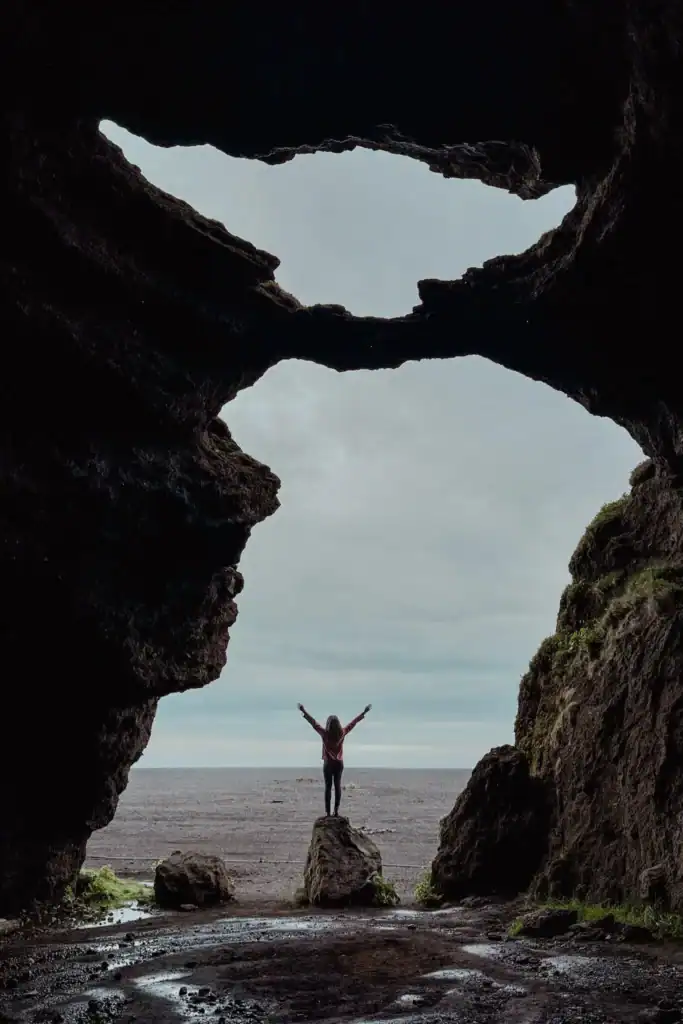 Person standing with arms raised inside a sea cave with large openings to the sky and ocean.