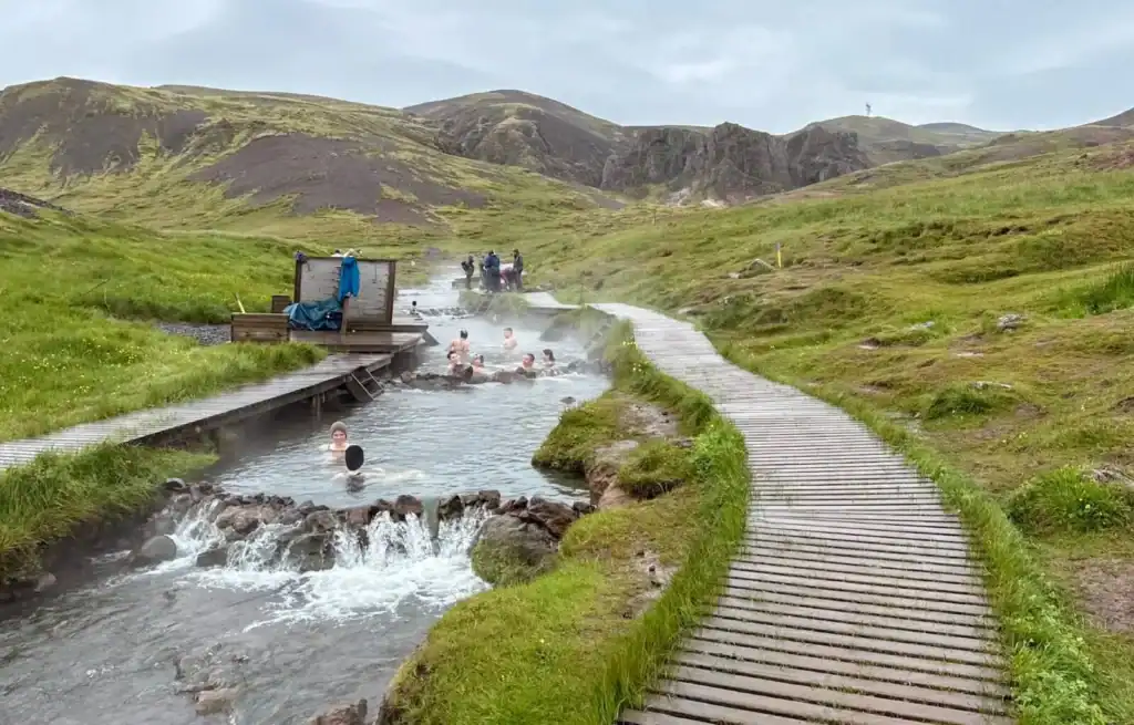 People relaxing in the geothermal river with a wooden walkway in a green, hilly landscape in Iceland.