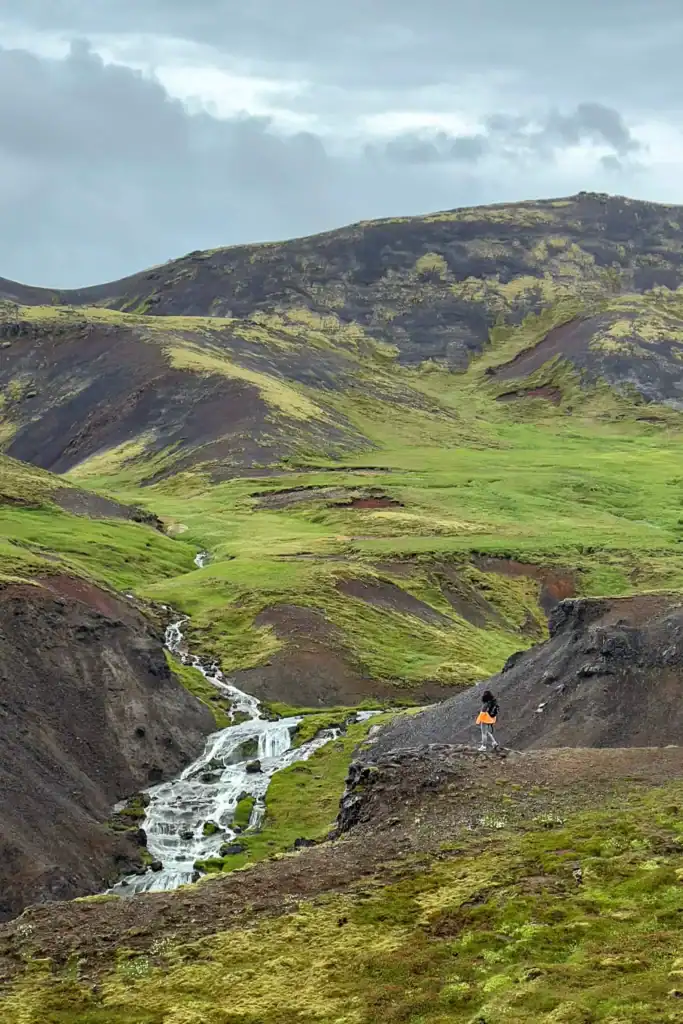 Person walking near a small waterfall in a green, hilly landscape with dark volcanic soil and cloudy sky.