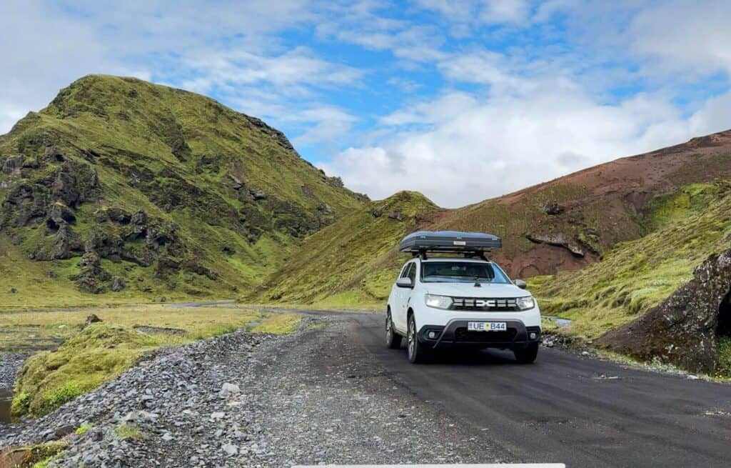 White SUV parked on a gravel road surrounded by green hills and mountains under a partly cloudy sky.