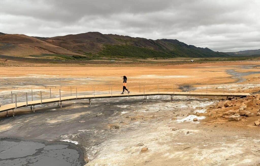 A person walking on a wooden bridge over geothermal mud pools in a volcanic landscape.