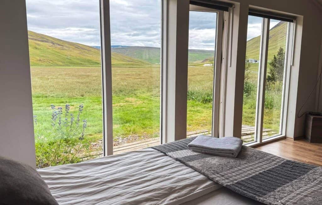 View of green hills and cloudy sky through large glass windows from a bedroom.