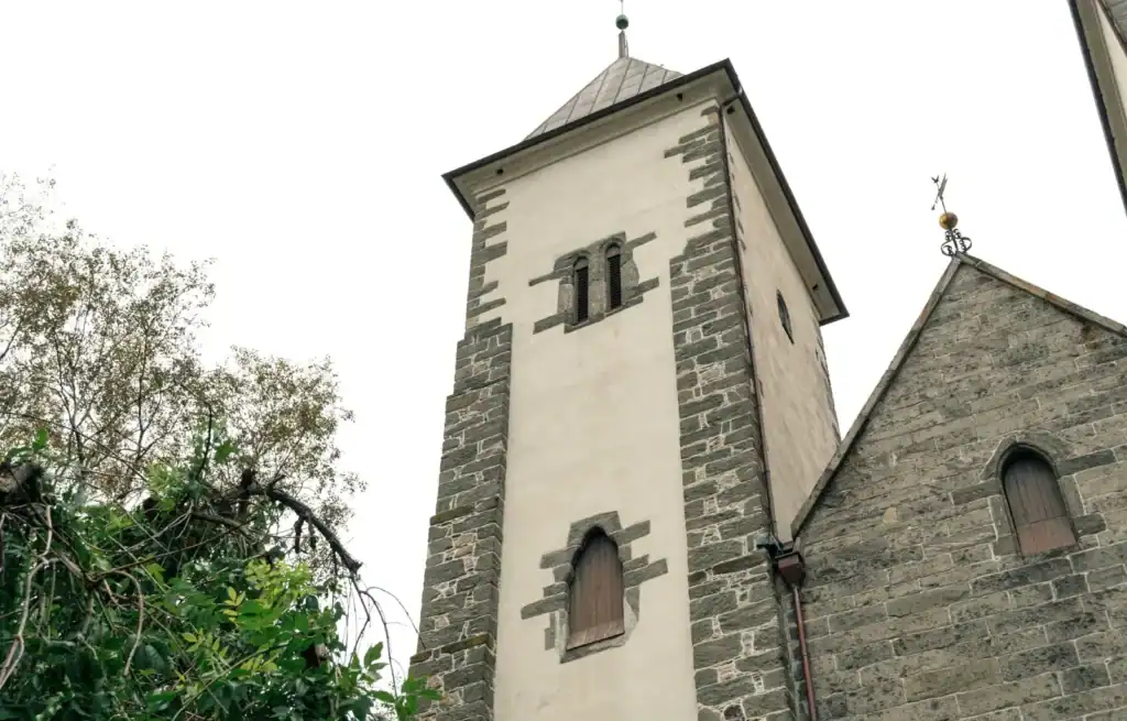 Historic stone church tower with small arched windows and a pointed roof, surrounded by trees.