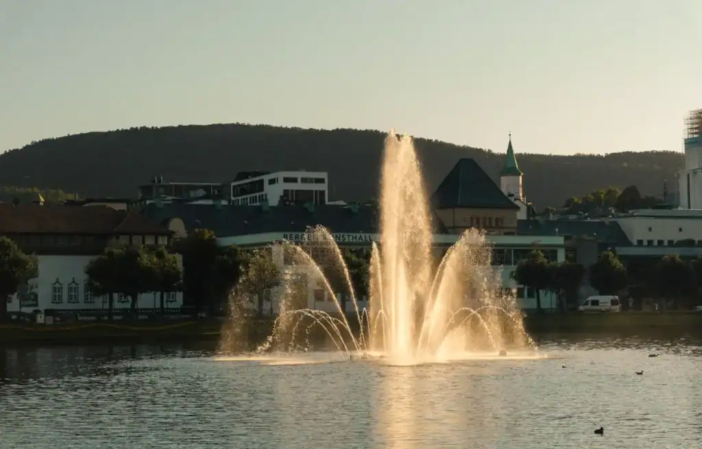 Fountain spraying water in a city park with buildings and hills in the background.