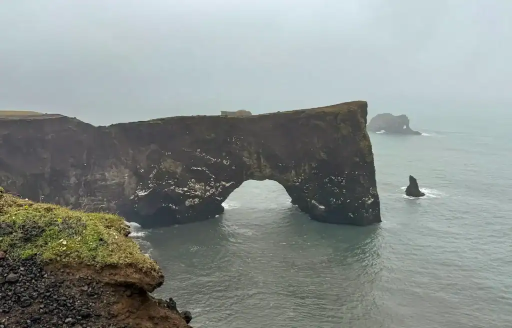 Cliffside arch formation on a foggy coastline with sea stacks in the distance.