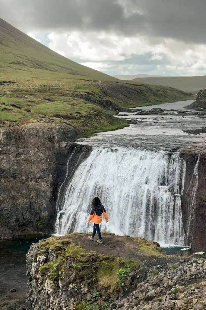 Person standing on mossy rock facing a waterfall in a green valley with cloudy sky.