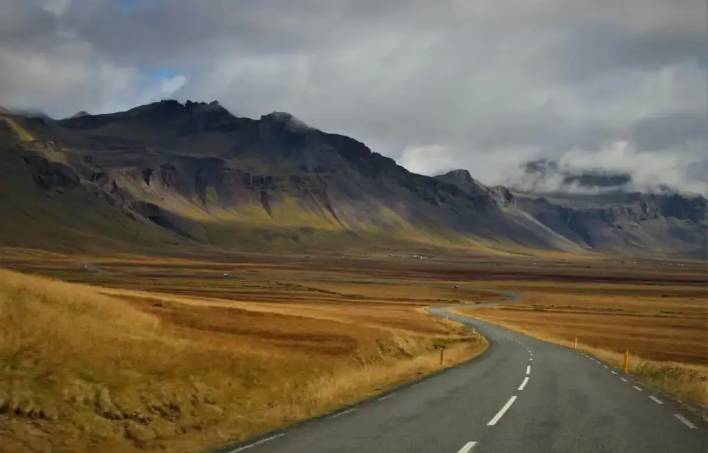 Winding road through vast Icelandic plains with mountains in the background under cloudy sky.