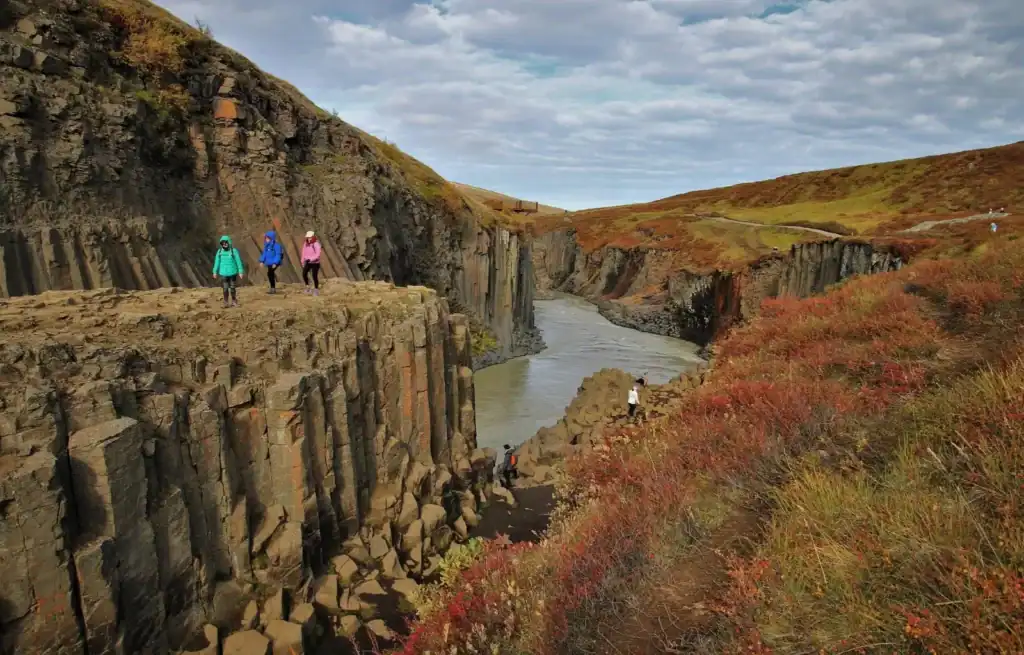Hikers walking along basalt cliffs overlooking a river in a rugged Icelandic landscape.