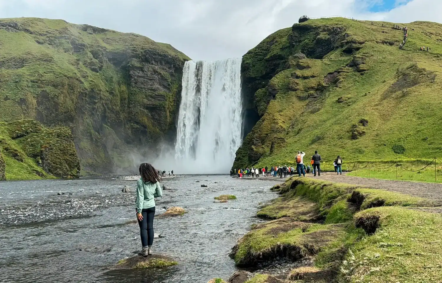 Person standing on rocks in a shallow river facing a large waterfall cascading between steep green cliffs, with visitors gathered along the walking path nearby. The lush landscape and mist rising from the falls create a dramatic natural scene