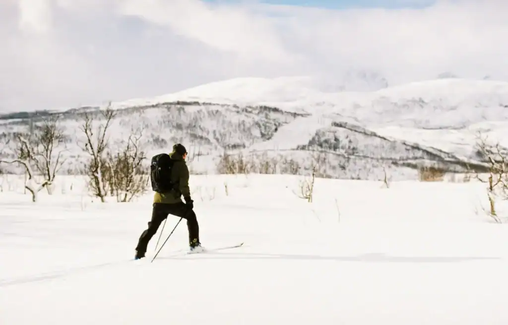 Person cross-country skiing through snow-covered landscape with hills and sparse trees in the background.