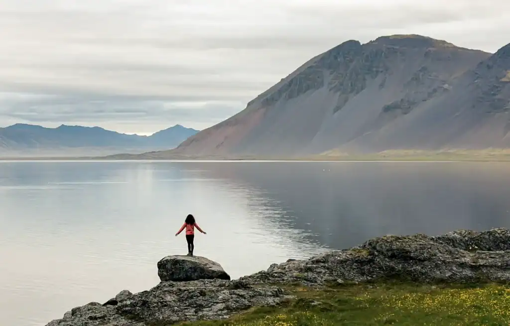 Person standing on a rock by a calm lake with mountains in the background under cloudy skies.